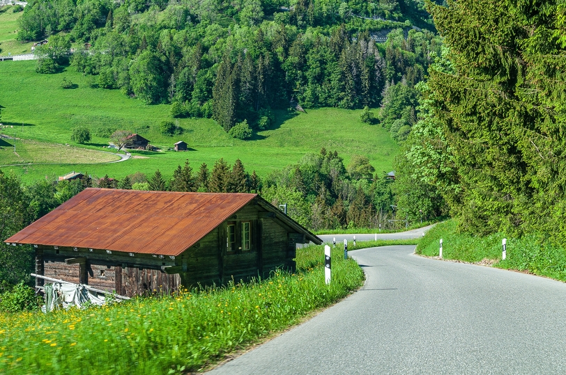 Landweg langs een houten huis in een groen heuvellandschap met bomen en weilanden.