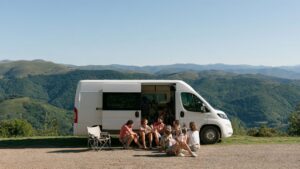 Groep vrienden zit voor een witte personenbus met uitzicht over groene bergen tijdens een roadtrip Spanje.